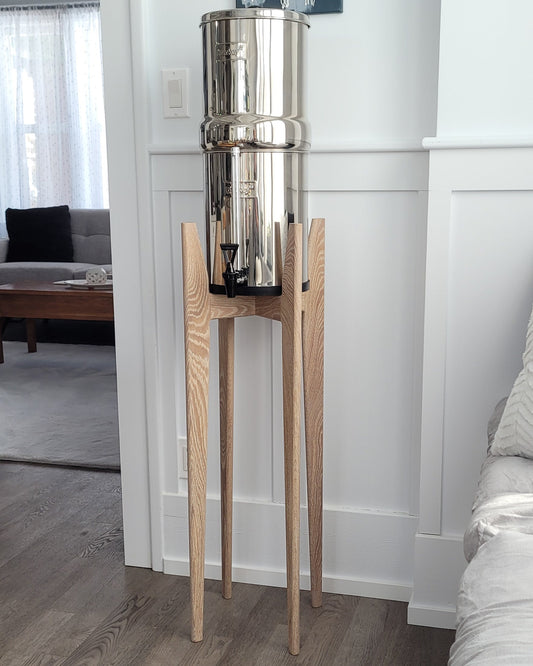 A beautiful white oak hardwood stand with tapered legs in a kitchen setting with a stainless steel Berkey water filter system standing on it . The stand is 38 inches tall in a mid century modern style with sloped upper posts for stability. the kitchen is a light colour