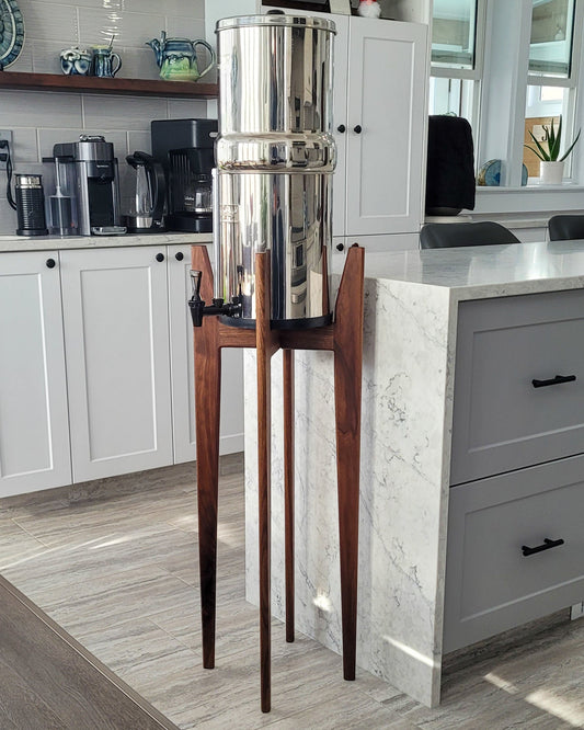 A beautiful walnut hardwood stand with tapered legs in a kitchen setting with a stainless steel Berkey water filter system standing on it . The stand is 38 inches tall in a mid century modern style with sloped upper posts for stability. the kitchen is a light colour