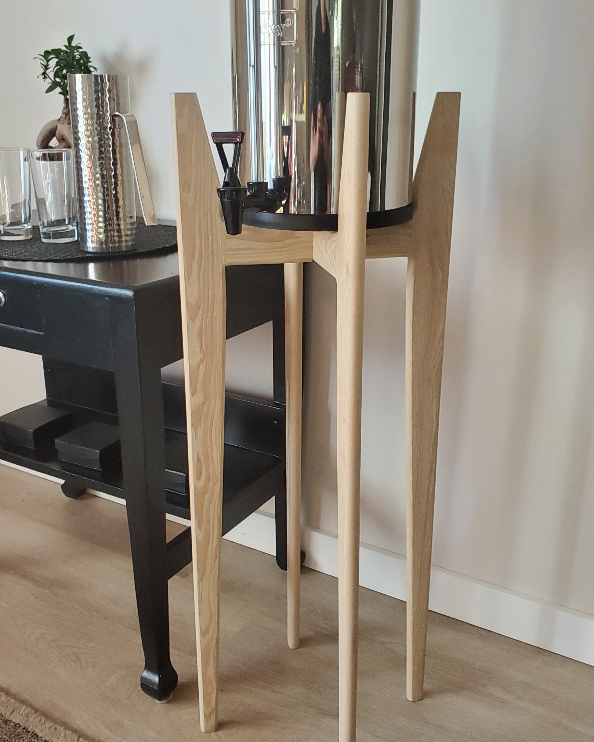 A solid Ash hardwood stand with a whitewash finish, designed to hold a water filter system, displayed next to a dark-colored table with a plant on it.