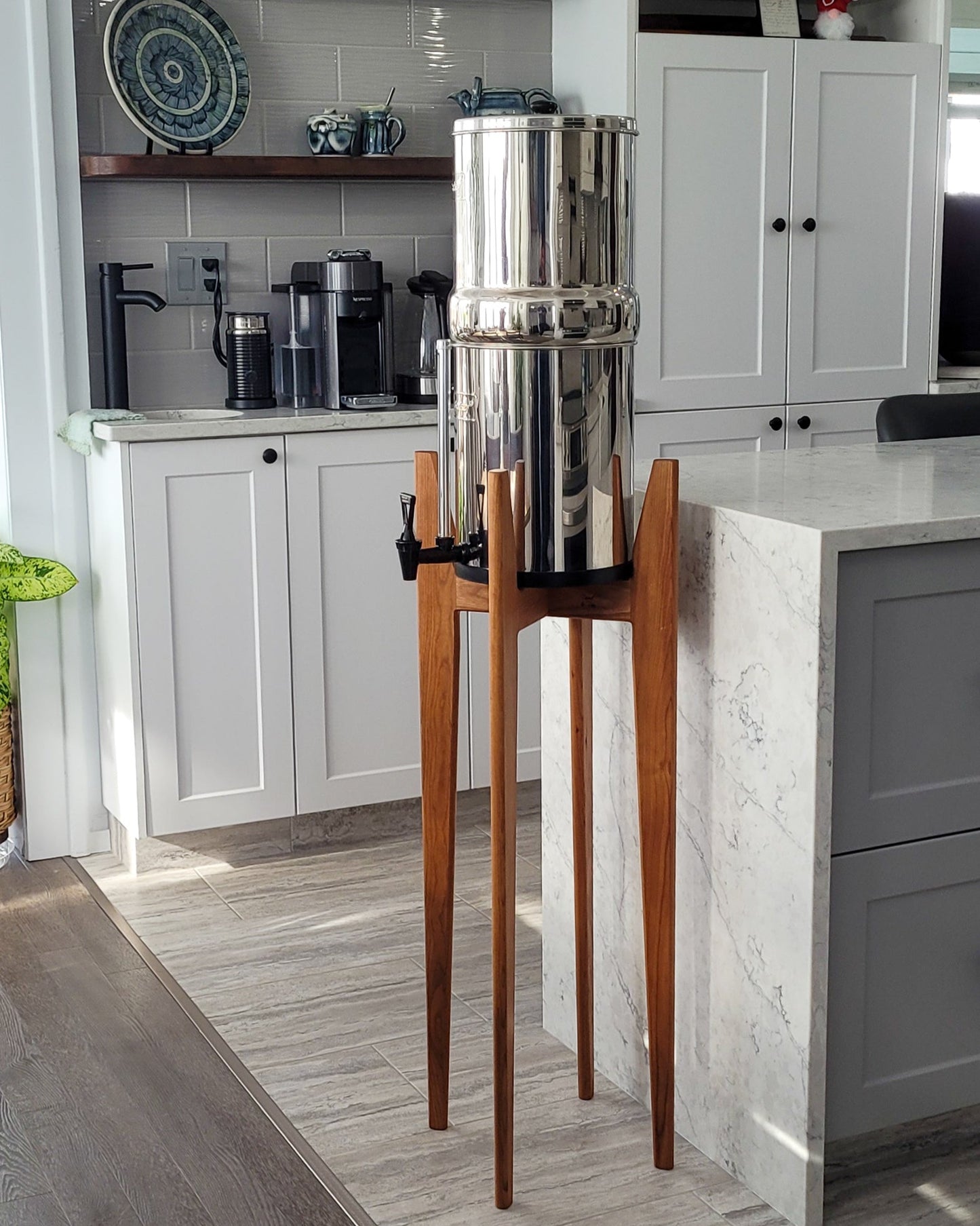 A beautiful cherry hardwood stand with tapered legs in a kitchen setting with a stainless steel Berkey water filter system standing on it . The stand is 38 inches tall in a mid century modern style with sloped upper posts for stability. the kitchen is a light colour