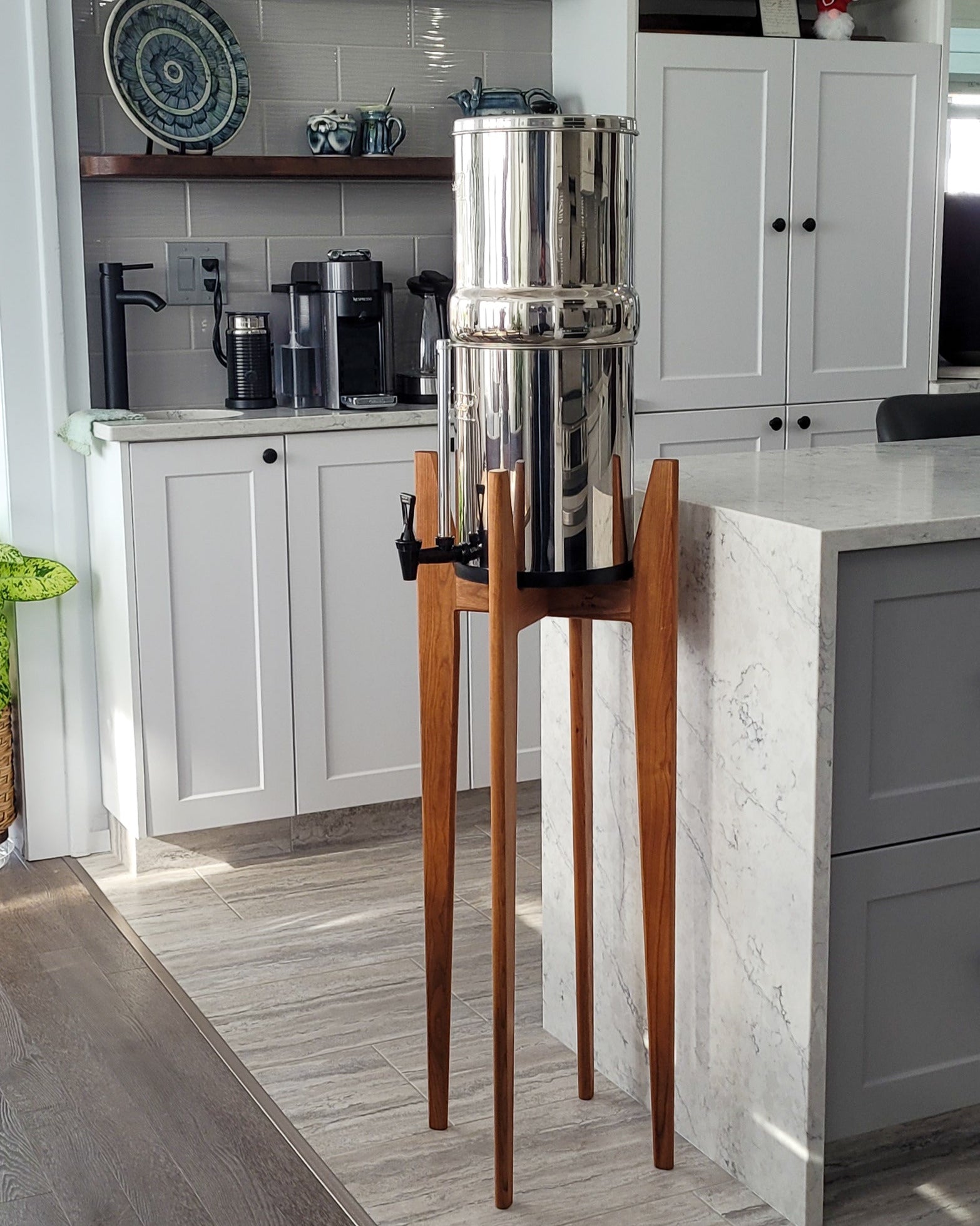 A beautiful cherry hardwood stand with tapered legs in a kitchen setting with a stainless steel Berkey water filter system standing on it . The stand is 38 inches tall in a mid century modern style with sloped upper posts for stability. the kitchen is a light colour