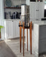 A beautiful cherry hardwood stand with tapered legs in a kitchen setting with a stainless steel Berkey water filter system standing on it . The stand is 38 inches tall in a mid century modern style with sloped upper posts for stability. the kitchen is a light colour