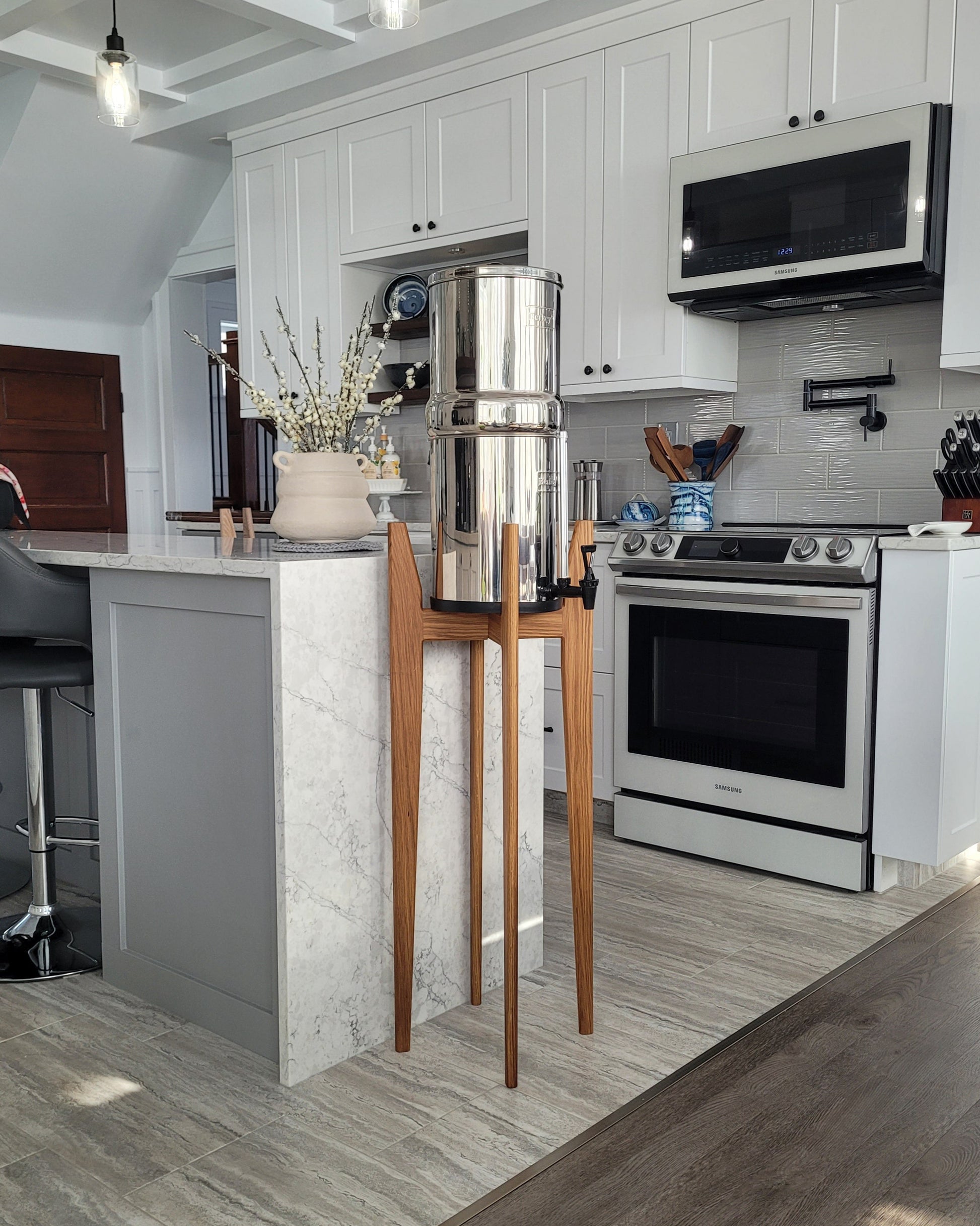A beautiful medium toned oak hardwood stand with tapered legs in a kitchen setting with a stainless steel Berkey water filter system standing on it . The stand is 38 inches tall in a mid century modern style with sloped upper posts for stability. the kitchen is a light colour