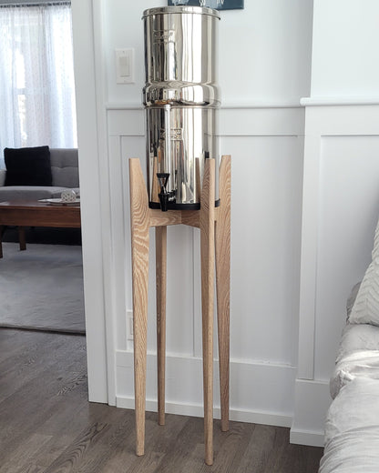A beautiful white oak  hardwood stand with tapered legs in a kitchen setting with a stainless steel Berkey water filter system standing on it . The stand is 38 inches tall in a mid century modern style with sloped upper posts for stability. the kitchen is a light colour