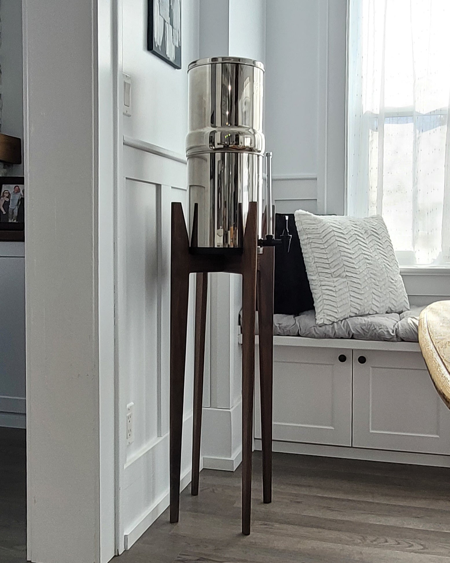 A beautiful walnut hardwood stand with tapered legs in a kitchen setting with a stainless steel Berkey water filter system standing on it . The stand is 38 inches tall in a mid century modern style with sloped upper posts for stability. the kitchen is a light colour